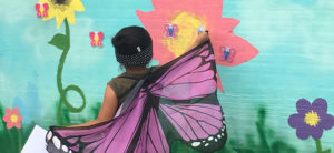 photo - girl wearing butterfly purple/black/white strap on wings, standing in front of wall painted with flowers and more decorative butterfly designs