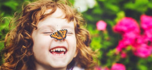 photo - laughing girl, with red hair, with monarch orange/black butterfly sitting on her nose, background has greenery and pink flowers