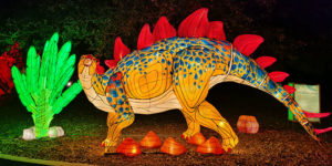 graphic - wild lites orange, tan with blue markings, long tail stegosaurus, has red spikes from neck to end of tail, standing among red rocks, green long leafed bush, with green bushes in background