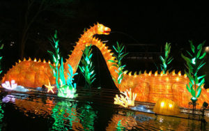photo - two wild lights dinosaurs, both orange color, with long necks, w/spikes running down from neck to end of tail. standing in pond, with long green bushes, and yellow bushes through out pond