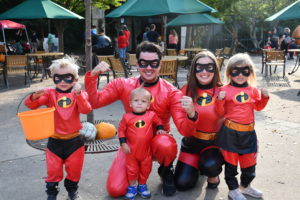 photo of family dressed as The Incredibles for halloween, wearing incredible red costumes, with "i" emblem on front, wearing black eye masks, posing on zoo plaza, tables, chairs, with umbrellas in background.