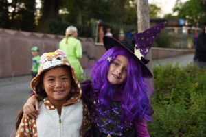 photo - two young girls, dressed; Halloween, one witch with purple long hair, witch's hat, with stars, purple shirt; other child wearing Sandy character costume, white polka dots on brown color, with hood that has face of character, girl has freckles on nose, cheeks.