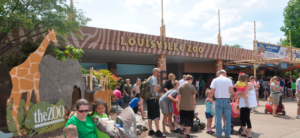 photo - large group of visitors with their families, on front plaza, during summer, with old louisville zoo signage on main entrance, with animal image display to left for photo opportunities.
