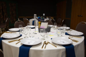 photo - event and venue set table with place settings for 8 guests, white plates, with gold silverware, blue napkins, 8 clear short stem glasses, 8 small clear class coffee cups, with blk basket with gifts inside, table number also