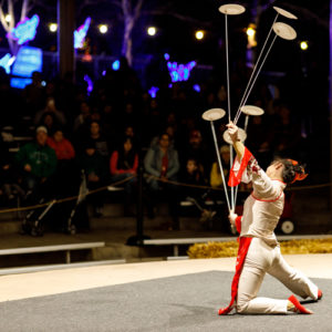 graphic - wild lights; graphic of girl in white costume, kneeling and balancing 5 plates on rods, for meta zoo audience, with array of lights hanging from ceiling