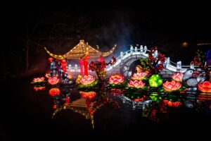 graphic - wild lights pond display of gray bridge, with variety of pink, green, orange, red, flowers throughout, with variety of colored birds orange and grey bluish, with a rack that holds the flowers, a red, gold pagoda, with pond reflections.