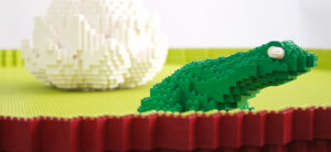 graphic - Lego sculpture of green frog and white lily pad on yellow Lego sculpture of a pond