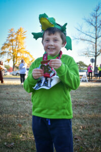 Child wearing alligator hat at Throo the Zoo 5K showing camera his runner's medal.