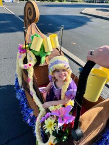 photo - female child dressed in purple dress, with yellow hat, w/yellow long braid, sitting in stroller decorated as brown boat, decorated with various odds, ends, flowers pushed by an adult