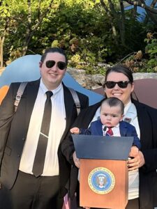 Family in front of the Zoo sign at Boo at the Zoo. Parents are dressed in suits as secret service. Mom has baby on front dressed as the "president" complete with a podium.