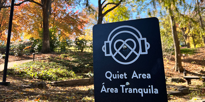photo - black sensory friendly sign w/white lettering, image of child with headphones on, also in spanish, park scenery in background