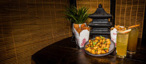 photo - wild lights display of different foods available, in chinese take out cartons, and on a plate, with 2 tall drinks with black pagoda in background