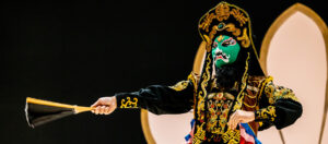 photo - wild lights facechanger character in black outfit with gold trim designs, embossed with other colorful designs of asian content, wearing green multi designed face features mask, holding a closed gold/black fan, background is black, with pink pedal stands